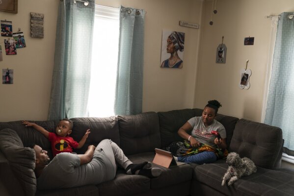 Darelyn Maldonado, 12, right, sits on the couch with her dog, Lisa, stepfather, Steven Depina, left, and 16-month-old brother, Elijah, at their home in Pawtucket, R.I., Wednesday, March 3, 2021. Maldonado, a seventh grade student, has been out of in person school for a year since the pandemic began. She feels like she's lost friends over the past year, has missed out on playing softball which she loves and just wants her life to go back to normal. (AP Photo/David Goldman)