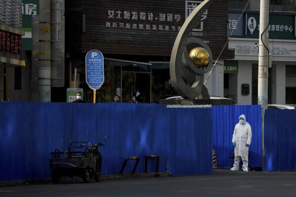 A worker in protective clothing keeps watch as residents wearing face masks stand near metal barriers set up around shuttered shop houses that were locked down as part of COVID-19 controls in Beijing on Thursday, Nov. 10, 2022. China's capital Beijing has closed city parks and imposed other restrictions as the country faces a new wave of COVID-19 cases. (AP Photo/Andy Wong)