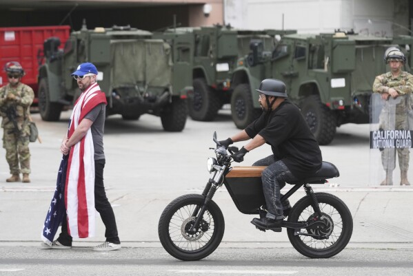 A lone demonstrator draped in a U.S. flag walks outside of the Metropolitan Detention Center Thursday, June 12, 2025, in Los Angeles. (AP Photo/Damian Dovarganes)