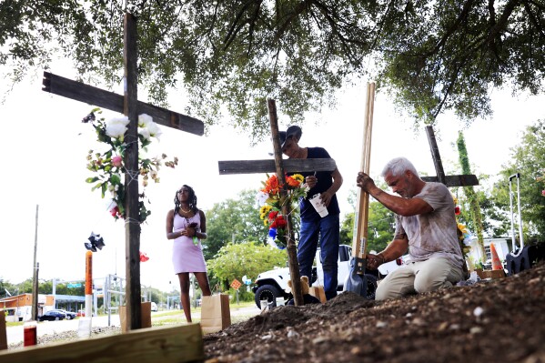 From left, a woman who goes by the name of "Queen," views crosses put up in memory of the victims of Saturday's shooting as artist Roberto Marquez, of Dallas, paints and Will Walsh, of Nocatee, Fla., helps construct posts Monday, Aug. 28, 2023, near the site of the attack at a Dollar General store in Jacksonville, Fla. Queen says she is a manager at the store and was holed up in the office at the store when the shooting occurred. (Corey Perrine/The Florida Times-Union via AP)