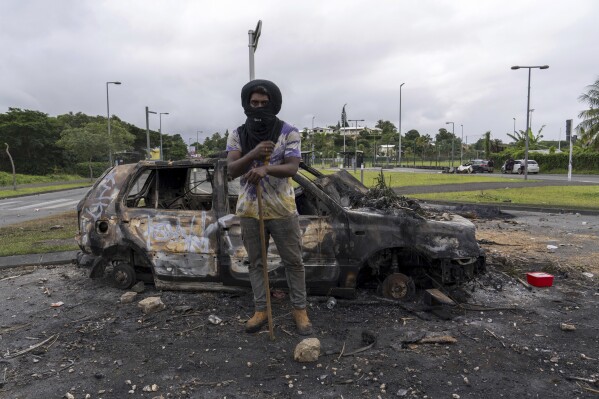 A man stands in front a burnt car after unrest in Noumea, New Caledonia, Wednesday May 15, 2024. France has imposed a state of emergency in the French Pacific territory of New Caledonia. The measures imposed on Wednesday for at least 12 days boost security forces' powers to quell deadly unrest that has left four people dead, erupting after protests over voting reforms. (AP Photo/Nicolas Job)