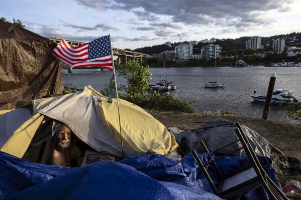 FILE - Frank, a homeless man sits in his tent with a river view in Portland, Ore., Saturday, June 5, 2021. The city council in Portland, Oregon, has approved new homeless camping rules. Under the rules, people who reject offers of shelter can face penalties, including fines of up to $100 or up to seven days in jail. (AP Photo/Paula Bronstein, File)
