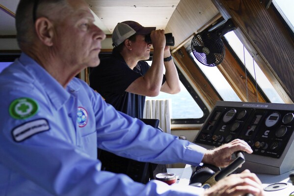 Aaron Smith, President and CEO of the Offshore Marine Service Association, center, peers through binoculars at ships installing the South Fork Wind project, as Capt. Rick Spaid, left, pilots the vessel Jones Act Enforcer, Tuesday, July 11, 2023, off the coast of Rhode Island. The trade association that represents the offshore service industry is going to great lengths to make sure that jobs go to Americans as the U.S. offshore wind industry ramps up. (AP Photo/Charles Krupa)