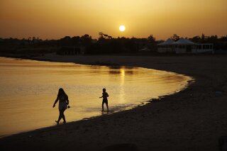 FILE - In this file photo dated Sunday, May 17, 2020, beachgoers walk in the sea water during sunset at an empty stretch of Dome beach at Makrinissos in Cyprus' seaside resort of Ayia Napa, a favorite among tourists.  Cyprus' government Wednesday May 27, 2020, is pledging to cover all costs for anyone testing positive for the coronavirus while on vacation in the east Mediterranean island nation, covering the costs of lodging, food, drink and medication for COVID-19 patients and their families.  (AP Photo/Petros Karadjias, FILE)