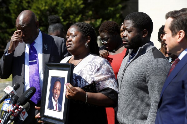 FILE - Caroline Ouko, mother of Irvo Otieno, holds a portrait of her son with attorney Ben Crump, left; her older son, Leon Ochieng; and attorney Mark Krudys, at the Dinwiddie Courthouse in Dinwiddie, Va., March 16, 2023. On Sunday, May 5, 2024, a Virginia judge signed off on a prosecutor's request to withdraw charges against five more people in connection with the 2023 death of Otieno, a young man who was pinned to the floor for about 11 minutes while being admitted to a state psychiatric hospital. (Daniel Sangjib Min/Richmond Times-Dispatch via AP, File)