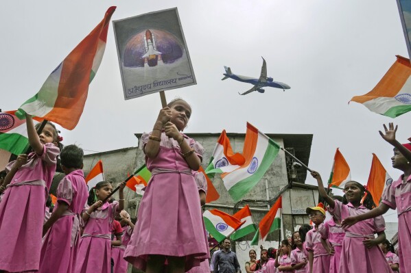  An aircraft prepares to land as children cheering for the successful landing of India's moon craft Chandrayaan-3, on the moon surface, wave Indian flags at a school near the airport in Mumbai, India, Tuesday, Aug.22, 2023. (AP Photo/ Rajanish Kakade, File)