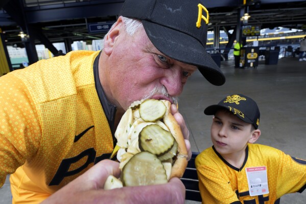 Tim Suwinski, father of Pittsburgh Pirates' Jack Suwinski, left, and grandson Will Hackl, both of Chicago, share a Renegade hot dog before a baseball game between the Pittsburgh Pirates and the Boston Red Sox in Pittsburgh, Friday, April 19, 2024. (AP Photo/Gene J. Puskar)