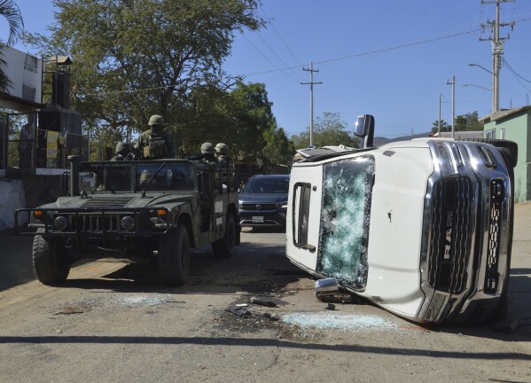 Army soldiers drive past a destroyed vehicle on the streets of Jesus Maria, Mexico, on Saturday, Jan. 7, 2023, the small town where Ovidio Guzman was detained earlier in the week. Thursday's government operation to detain Ovidio, the son of imprisoned drug lord Joaquin "El Chapo" Guzman, unleashed firefights that killed 10 military personnel and 19 suspected members of the Sinaloa drug cartel, according to authorities. (AP Photo/Martin Urista)