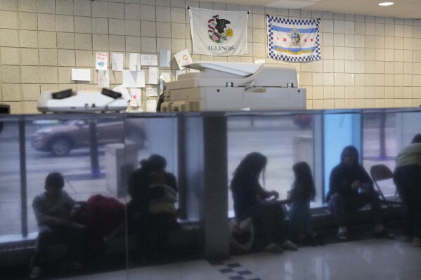 FILE - Immigrants from Venezuela are reflected in a marble wall while taking shelter at the Chicago Police Department's 16th District station on Monday, May 1, 2023. In Chicago, where 13,000 migrants have settled in the last year, Mayor Brandon Johnson and Illinois Gov. J.B. Pritzker wrote Homeland Security Secretary Alejandro Mayorkas to ask for parole for asylum-seekers, which, they say, would allow him to get around the wait for a work permit. (AP Photo/Charles Rex Arbogast, FIle)