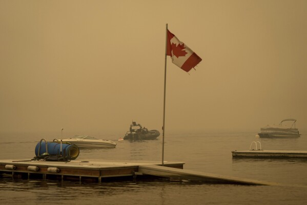 Thick smoke from the Lower East Adams Lake wildfire fills the air around a Canadian flag fluttering in the wind as Royal Canadian Mounted Police officers on a boat patrol Shuswap Lake, in Scotch Creek, British Columbia, on Sunday, Aug. 20, 2023. (Darryl Dyck/The Canadian Press via AP)