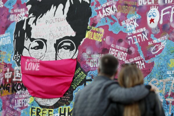 FILE - A couple look at the "Lennon Wall" with a face mask attached to the image of John Lennon, in Prague, Czech Republic, on April 6, 2020. Like so many other events in the year of coronavirus, a...