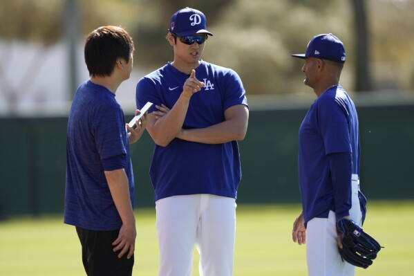 Los Angeles Dodgers' Shohei Ohtani, center, with interpreter Ippei Mizuhara, left, talks with coach Dino Ebel, right, during spring training baseball workouts at Camelback Ranch in Phoenix, Sunday, March 3, 2024. (AP Photo/Carolyn Kaster)