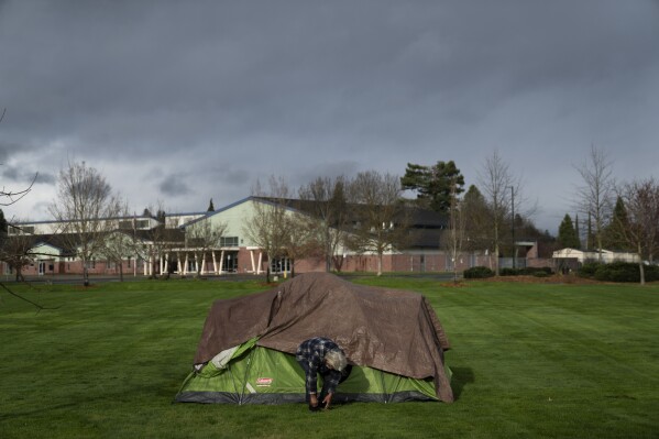 With Fruitdale Elementary School in the background, a homeless man adjusts his shoe at Fruitdale Park, March 23, 2024, in Grants Pass, Ore. (AP Photo/Jenny Kane, File)