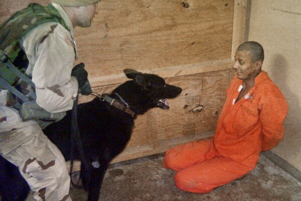 This undated still photo made available by The Washington Post on May 21, 2004, shows a U.S. soldier, who was identified in a military court-martial as Sgt. Michael J. Smith, holding a dog in front an Iraqi detainee at Abu Ghraib prison on the outskirts of Baghdad. A federal judge has again refused to dismiss a lawsuit brought by former Abu Ghraib inmates against a military contractor they accuse of being complicit in torture at the infamous Iraqi prison. The horrific mistreatment of prisoners there two decades ago sparked international outrage when photos became public of smiling U.S. soldiers posing in front of abused prisoners. (The Washington Post via AP)