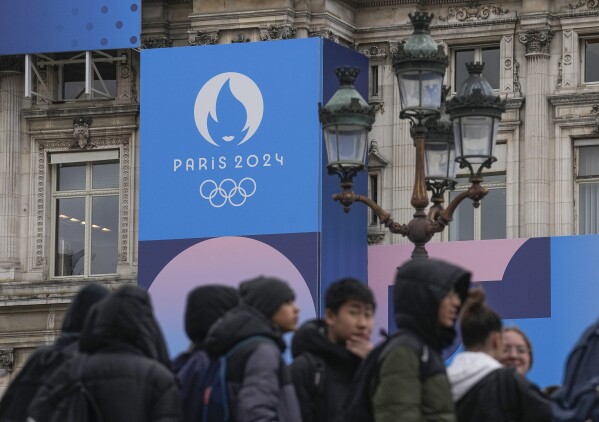Youth gather at Paris City Hall where a Paris 2024 Olympic sign is displayed in Paris, Tuesday, March 5, 2024. The French government announced that tourists won't be allowed to watch the opening ceremony of the Paris Olympics for free as initially promised, the government announced Tuesday, as it grapples with security concerns about the unprecedented open-air event along the Seine River. (AP Photo/Michel Euler)