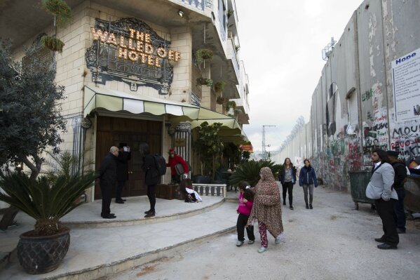 FILE - in this Friday, March 3, 2017 file photo, People pass by the "The Walled Off Hotel" and the Israeli security barrier the West Bank city of Bethlehem. As visitors descend on Bethlehem this Christmas, they have the option of staying in the restored centuries-old guesthouses, taking food tours of local markets and perusing the dystopian art in and around a hotel designed by the British graffiti artist Banksy. (AP Photo/Dusan Vranic, File)
