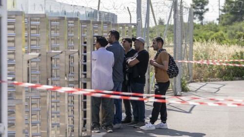 A group of men wait at the turnstiles and speak with survivors, mostly from Pakistan, of a deadly migrant boat sinking at a migrant camp in Malakasa north of Athens, on Monday, June 19, 2023. Hundreds of migrants are believed to be missing after a fishing trawler sank off southern Greece last week. (AP Photo/Petros Giannakouris)