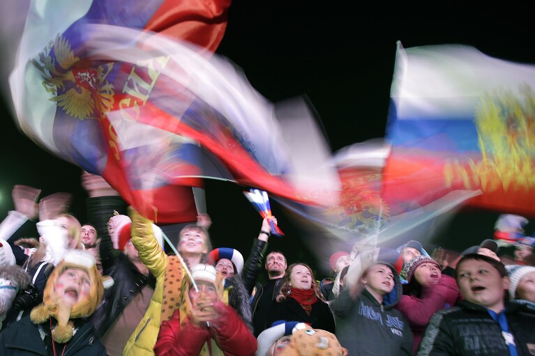 FILE - Russian families gather and wave the flag during the national anthem as they watch the telecast of the opening ceremony of the 2014 Winter Olympics in Sochi, Russia, on Feb. 7, 2014. It cost Russia an estimated $55 billion to prepare and host the Olympics in the balmy Black Sea resort of Sochi, where most facilities, including 11 sports venues, were built from scratch along with highways, rail lines and other infrastructure for athletes and spectators. (AP Photo/Wong Maye-E, File)