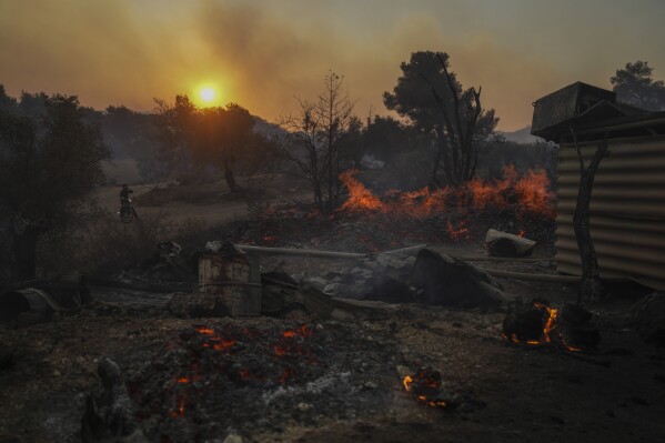 A man on a motorcycle looks on as a shipyard which is on fire in Mandra west of Athens, on Tuesday, July 18, 2023. In Greece, where a second heatwave is expected to hit Thursday, three large wildfires burned outside Athens for a second day. Thousands of people evacuated from coastal areas south of the capital returned to their homes Tuesday when a fire finally receded after they spent the night on beaches, hotels and public facilities. (AP Photo/Petros Giannakouris)