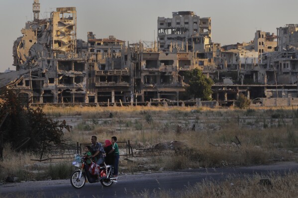 FILE - People ride their motorcycle by damaged buildings in the old town of Homs, Syria, Aug. 15, 2018. Syria’s civil war has entered its 14th year on Friday March 15, 2024, a somber anniversary in a long-frozen conflict. The country is effectively carved up into areas controlled by the Damascus government, various opposition groups and Kurdish forces. (AP Photo/Sergei Grits, File)