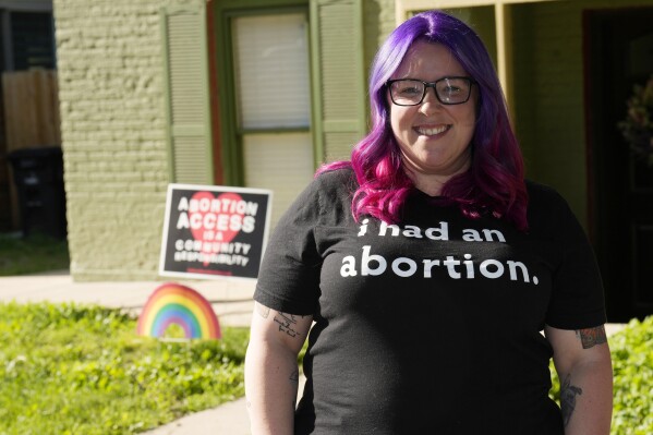 Leah Dean, a native of the Texas Panhandle, poses outside her home Monday, July 3, 2023, in Denver. Americans are segregating by their politics at a rapid clip, helping fuel the greatest divide between the states in modern history. In Texas, Dean had been scared to fly an abortion rights banner outside her house. Now in Denver, an LGBTQ+ pride flag flies above the banner in front of her house that proclaims, “Abortion access is a community responsibility.” (AP Photo/David Zalubowski)