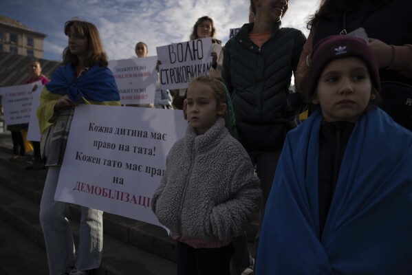 Relatives of soldiers hold a poster reading "Each kid has a right to have father, each father should have right for demobilization", while attending a meeting in Independence square in Kyiv, Ukraine, Friday, Oct. 27, 2023. Families of Ukrainian servicemen gathered to demand their loved ones return home after serving 18 months in the military as the war with Russia nears its second year. (AP Photo/Alex Babenko)