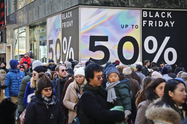 File - Crowds walk past a large store sign displaying a Black Friday discount in midtown Manhattan, Friday, Nov. 23, 2018, in New York. While Black Friday may no longer look like the crowd-filled, in-person mayhem that it was just decades ago — in large part due to the rising dependence on online shopping that was accelerated by the COVID-19 pandemic — the holiday sales event is still slated to attract millions of consumers. (AP Photo/Bebeto Matthews, File)