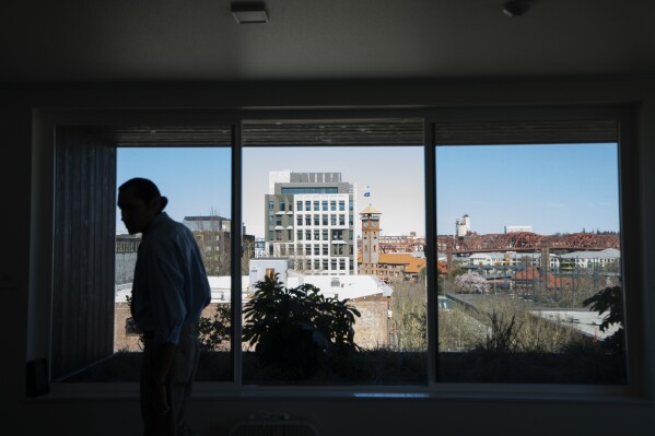 With a view of Portland, Ore., in the background, Cheyenne Welbourne walks in the common room at The Starlight affordable housing building that is run by Central City Concern, a Portland-based homeless services nonprofit, on Friday, March 15, 2024. Welbourne moved into one of the nonprofit's single room occupancy units in downtown Portland last March after years of living on the streets. (AP Photo/Jenny Kane)