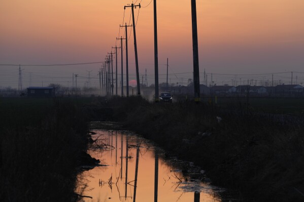 The sun sets near pylons as an electric vehicle passes a dirt road in the rural outskirts of Weifang in eastern China's Shandong province on March 22, 2024. Chinese battery companies, EV manufacturers and utilities are all racing to develop more advanced batteries to store the electricity from solar panels. (AP Photo/Ng Han Guan)
