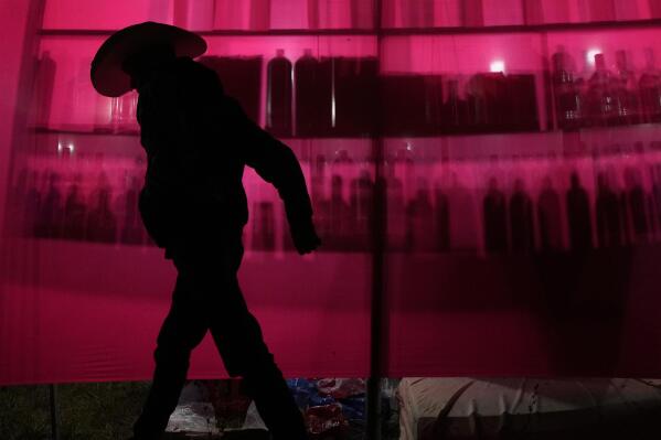 A man walks past liquor bottles silhouetted against a red cloth during the grand dance of the religious tradition, "Folia do Divino Espirito Santo" or Feast of the Divine, in the rural area of Pirenopolis, state of Goias, Brazil, Saturday, May 28, 2022. The event celebrates the coming of the Holy Spirit to Jesus' apostles after his crucifixion and has been performed for two centuries since it was first brought to Brazil by Portuguese colonizers. Once there, it was influenced by the cultures of Indigenous people and Black slaves. (AP Photo/Eraldo Peres)