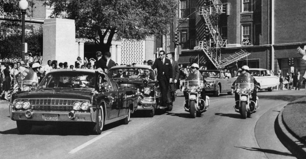 FILE - Seen through the foreground convertible's windshield, President John F. Kennedy's hand reaches toward his head within seconds of being fatally shot, as first lady Jacqueline Kennedy holds his forearm, as the motorcade proceeds along Elm Street past the Texas School Book Depository, Nov. 22, 1963, in Dallas. The 60th anniversary of President Kennedy's assassination, marked on Wednesday, Nov. 22, 2023, finds his family, and the country, at a moment many would not have imagined in JFK's lifetime. (AP Photo/James W. "Ike" Altgens, File)