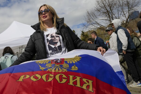 A woman wearing a shirt with a picture of Russian President Vladimir Putin holds a Russian flag outside a polling station set up in a Russian school in Belgrade, Serbia, Sunday, March 17, 2024. Russians at home and abroad headed to the polls for a presidential election that was all but certain to extend President Vladimir Putin's rule after he clamped down on dissent. (AP Photo/Darko Vojinovic)