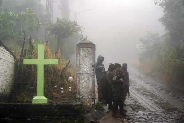 Soldiers stand guard under the rain near a cross that decorates a road in Comasagua, El Salvador, Monday, Oct. 10, 2022. At least five soldiers died on Monday nearby when the wall of a home collapsed as they were sheltering from the rain while deployed to crackdown on gang activity. (AP Photo/Moises Castillo)