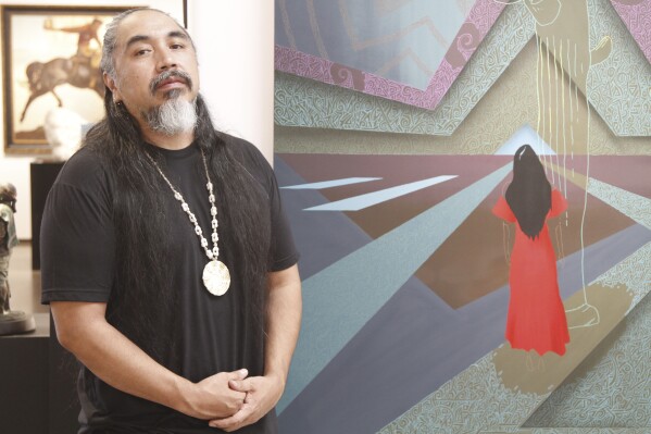 Artist Thomas "Breeze" Marcus attends an exhibition opening of his work at Blue Rain Gallery in Santa Fe, N.M. on Thursday, Aug. 17, 2023, including the painting titled "Answered Prayer," backgound. A separate group exhibition that included his work was postponed in Arizona amid outrage over a painting by Shepard Fairey that is critical of police. It's the latest of several recent incidents around the U.S. where artists in public spaces have had the rug pulled out from under them because of the content of their work. (AP Photo/Morgan Lee)