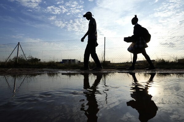 A man and woman walks over puddled water along a street from an overflowing reservoir in Hamanskraal, Pretoria, South Africa, Friday, May 26, 2023. Health authorities are yet to confirm the exact source of the cholera outbreak, but poor waste water management and local government instability in South Africa's capital city have been blamed for the situation. (AP Photo/Themba Hadebe)
