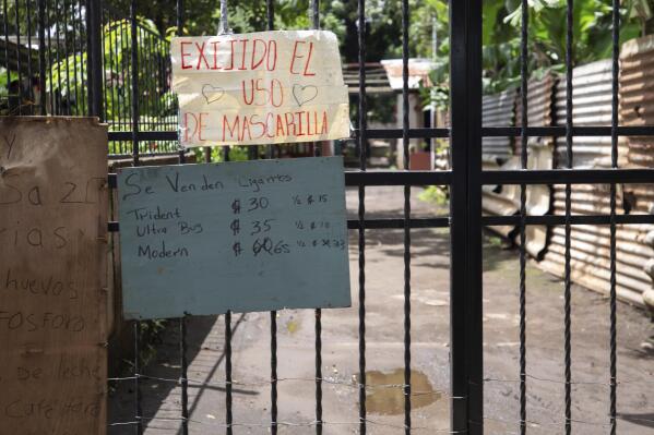 A sign requiring the use of masks during the COVID-19 pandemic hangs over signs advertising cigarettes, eggs, matches, milk and coffee for sale on a gate at the entrance to a residential area in Managua, Nicaragua, Thursday, Sept. 9, 2021. (AP Photo/Miguel Andrés)