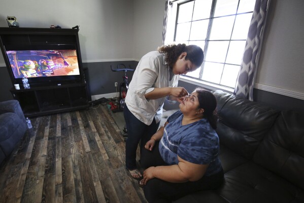 Marvin Estela Pineda, right, originally from El Salvador and blind from glaucoma, gets eye drops from her daughter Mayde at their home in Madera, Calif., Thursday, May 30, 2024. California Gov. Gavin Newsom is facing criticism for his proposal to eliminate an optional Medicaid benefit for some disabled immigrants. (AP Photo/Gary Kazanjian)