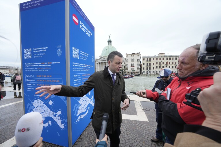 Членът на съвета на Венеция Симоне Вентурини говори с репортери пред тотема за туристически данък във Венеция, Италия, сряда, 24 април 2024 г. Плажният град Венеция започва пилотна програма в четвъртък, 25 април 2024 г., за да таксува входна такса от 5 евро на еднодневни екскурзии, които властите се надяват да обезсърчат туристите да пристигат в дните на пика.  Длъжностните лица очакват около 10 000 души да платят входни такси в града през първия ден, като изтеглят QR код, за да докажат плащането си.  (AP Photo/Лука Бруно)
