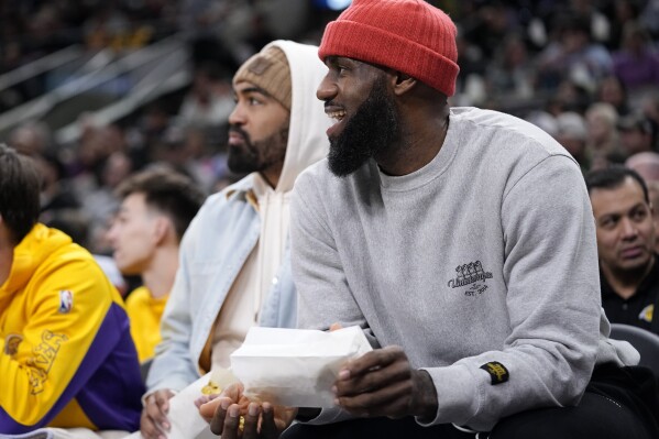 Los Angeles Lakers forward LeBron James, right, eats popcorn as he watches from the bench during the first half of an NBA basketball game against the San Antonio Spurs in San Antonio, Wednesday, Dec. 13, 2023. (AP Photo/Eric Gay)