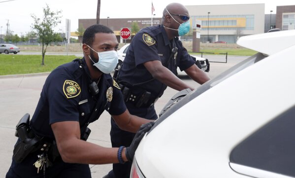 In this April 9, 2020, photo, amid concerns of the spread of COVID-19, Dallas ISD police officers Mylon Taylor, left, and Gary Pierre push a car that ran out of gas while waiting in line for the weekly school meal pick up for students in Dallas. The coronavirus pandemic that has crippled big-box retailers and mom and pop shops worldwide may be making a dent in illicit business, too. (AP Photo/LM Otero)