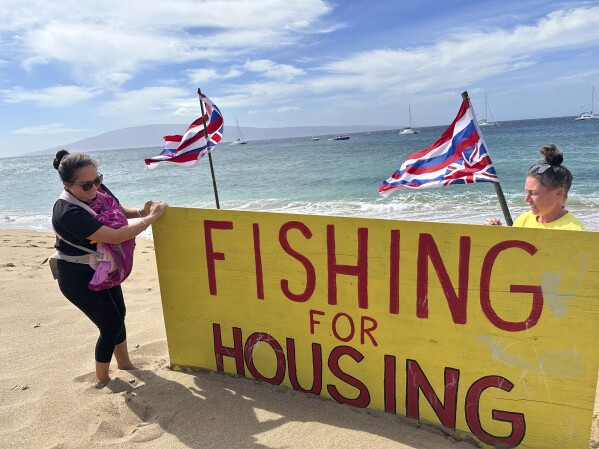 Jordan Ruidas, left, and Katie Austin adjust a sign on Kaanapali Beach in Lahaina, Hawaii on Wednesday, Nov.14, 2023. A group of Lahaina wildfire survivors is vowing to camp on a popular resort beach until the mayor uses his emergency powers to shut down unpermitted vacation rentals and make the properties available for residents in desperate need of housing. (AP Photo/Audrey McAvoy)