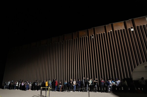 People line up against a border wall as they wait to apply for asylum after crossing the border from Mexico Tuesday, July 11, 2023, near Yuma, Arizona. (AP Photo/Gregory Bull)