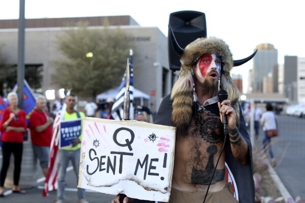 FILE - Jacob Anthony Chansley, who also goes by the name Jake Angeli, a Qanon believer speaks to a crowd of President Donald Trump supporters outside of the Maricopa County Recorder's Office where votes in the general election are being counted, in Phoenix on Nov. 5, 2020. From the Salem witch trials to fears of the Illuminati to the Red Scare to QAnon, conspiracy theories have always served as dark counter programming to the American story taught in history books. (AP Photo/Dario Lopez-Mills, File)
