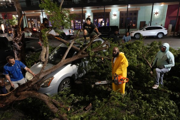 Rapper Trae tha Truth, in yellow, cuts fallen tree limbs on top of a car in the aftermath of a severe thunderstorm that passed through downtown, Thursday, May 16, 2024, in Houston. (AP Photo/David J. Phillip)