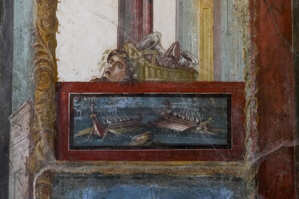 A detail of a fresco showing a sea battle framed in Pompeii red topped by a basket and a mask that decorates the living room of house of the Vetti, in one of the wealthiest homes in Pompeii, the House of the Vetti, in the Pompeii archeological site, Thursday, Dec. 7, 2023. A new project in Pompeii is combining traditional dyeing techniques with colorful frescoes, a historic reconstruction of a garden with Roman-era plants and the study of a steamy workshop where slaves dyed cloth, to revive the daily life in the ancient city before the eruption. (AP Photo/Andrew Medichini)