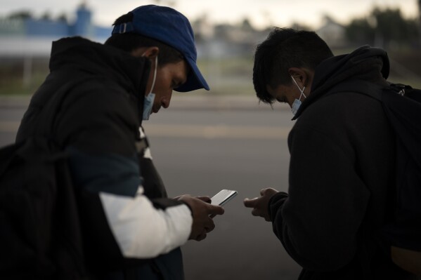 Migrants check their phones as they arrive at a bus stop after leaving a processing facility, Friday, Feb. 23, 2024, in San Diego. Hundreds of migrants were dropped off Friday at a sidewalk bus stop amid office parks in San Diego with notices to appear in immigration court after local government funding for a reception center ran out of money sooner than expected. (AP Photo/Gregory Bull)