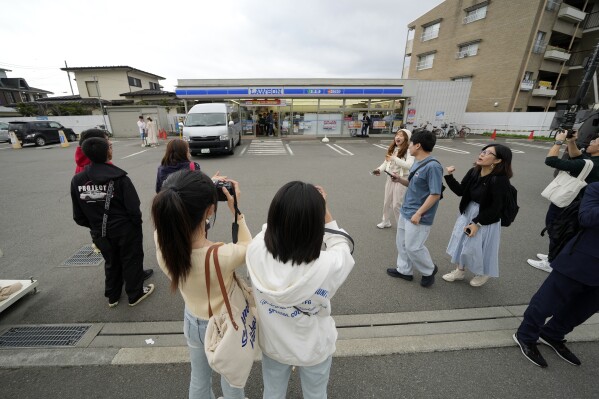 Touristen machen an einem bewölkten Abend am Dienstag, dem 30. April 2024, in der Stadt Fujikawaguchiko, Präfektur Yamanashi, Zentraljapan, Fotos vor dem Lawson Convenience Store, einem beliebten Fotospot mit atemberaubender Aussicht auf den Berg Fuji im Hintergrund.  Die Stadt Fujikawaguchiko, die für eine Reihe beliebter Drehorte für die japanische Marke „Mount Fuji“ bekannt ist, hat am Dienstag damit begonnen, eine riesige schwarze Leinwand auf dem Bürgersteig zu platzieren, um den Blick auf den Berg in einem Viertel zu versperren, das von Japans jüngstem Fall von Übertourismus heimgesucht wurde.  (AP Photo/Eugene Hoshiko)