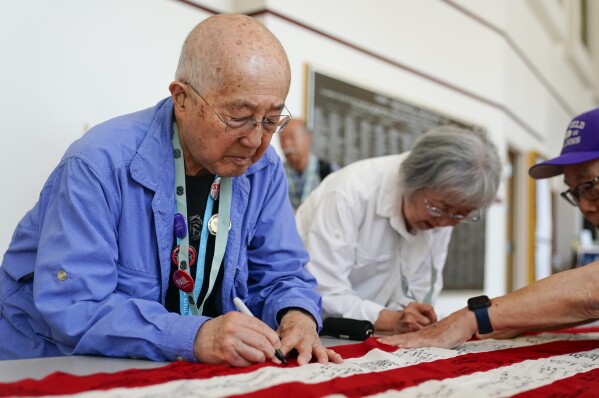 Minidoka survivor Paul Tomita, left, and wife Mabel Tomita, right, a survivor of Tule Lake and Gila River, sign a World War II-era 48-star flag as part of a project by Judge Johnny Gogo of Santa Clara County Superior Court at the College of Southern Idaho, Friday, July 7, 2023, in Twin Falls, Idaho. Gogo originally started the project to promote Fred Korematsu Day and has traveled around the country gathering signatures from survivors, filling several flags he has donated to Japanese American museums. (AP Photo/Lindsey Wasson)
