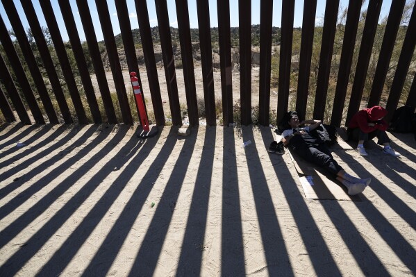 Two men from China rest in the partial shade of the border wall that separates Mexico from the United States after crossing to seek asylum, Tuesday, Oct. 24, 2023, near Jacumba, Calif. A major influx of Chinese migration to the United States on a relatively new and perilous route through Panama's Darién Gap jungle has become increasingly popular thanks to social media. (AP Photo/Gregory Bull)