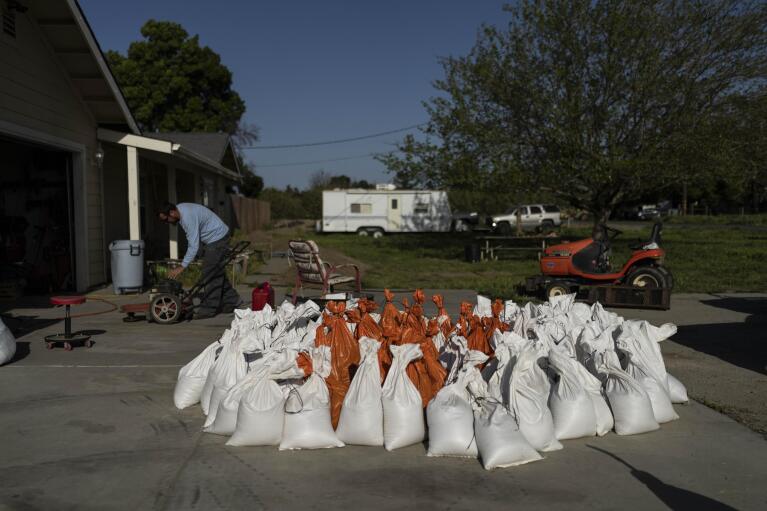 Brennon Williams operates a lawn mower at his girlfriend's home as sandbags are prepared in anticipation of flooding of the Kings River in the Island District of Lemoore, Calif., Wednesday, April 19, 2023. Residents in rural communities in the heart of the state are facing the prospect of being marooned by rising rivers or flooded out. (AP Photo/Jae C. Hong)